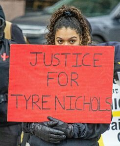 A woman holding a red sign that says “Justice for Tyre Nichols” in capital black letters.