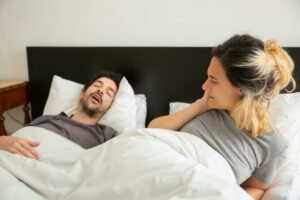 A man and woman in bed together. The man sleeps with his mouth open, and the woman is sitting up looking at him.