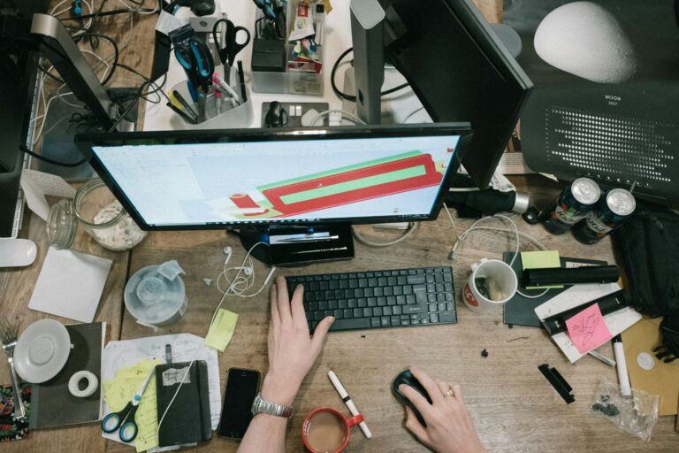 A crowded desk space with a desktop computer, office supplies, and drink cups.