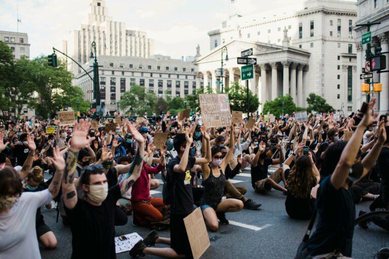 A large peaceful protest of people sitting in the street with signs and their hands up in the air.